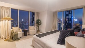 Ein modernes Hotelzimmer mit einem großen Bett, zwei Sesseln, einem kleinen Beistelltisch und Blick auf eine nächtliche Skyline.