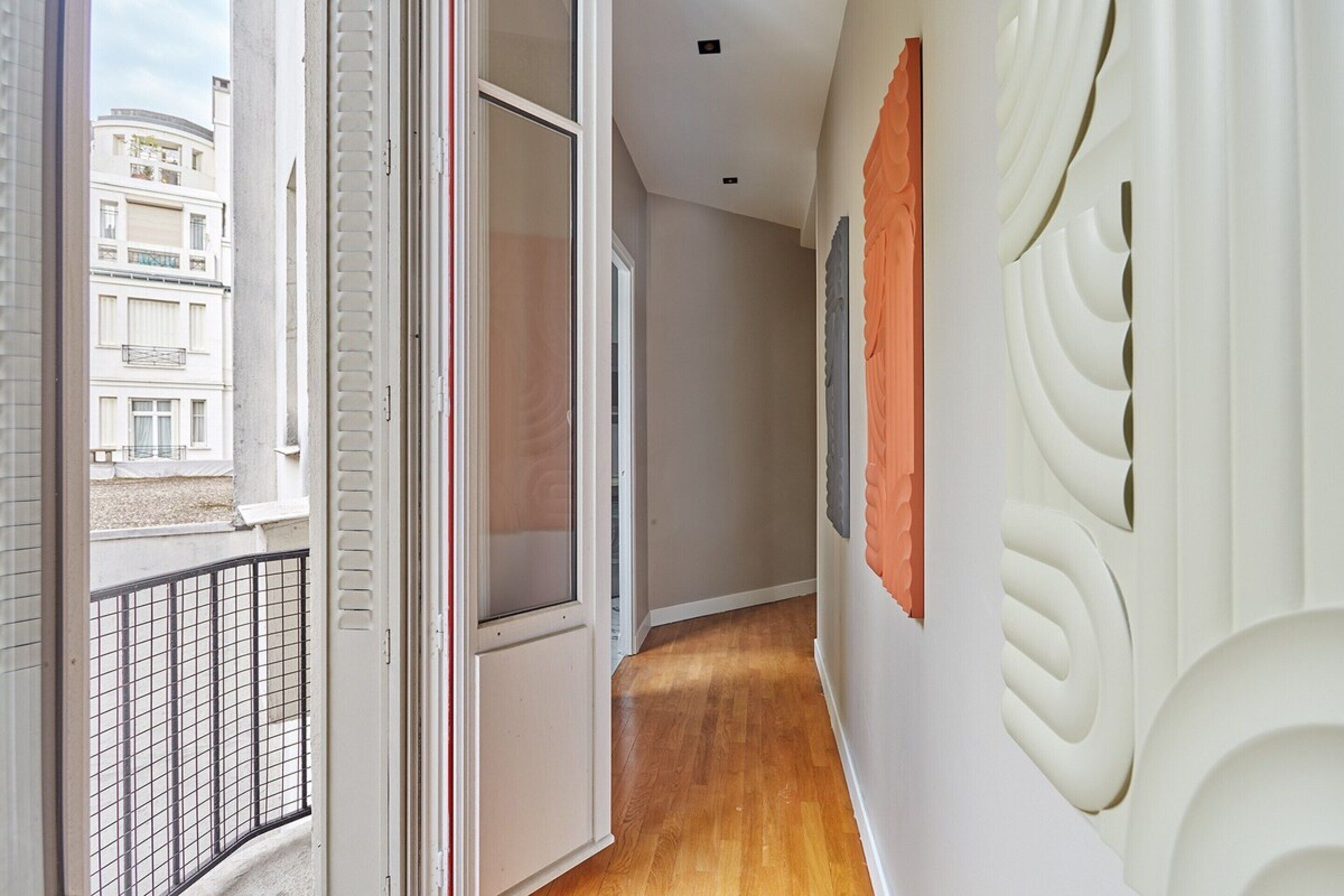A hallway with a wooden floor, a white wall with a decorative panel, and a door leading to another room.