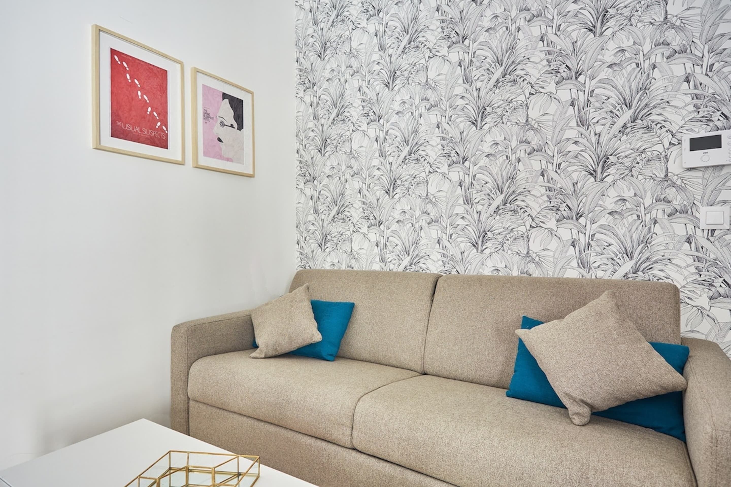 Un sofá color beige con cojines azules, una mesita de centro blanca y una pared con estampado floral.