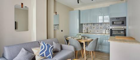 A modern living room with a grey sofa, a dining table with chairs, and a kitchen area with light blue cabinets and a built-in oven.
