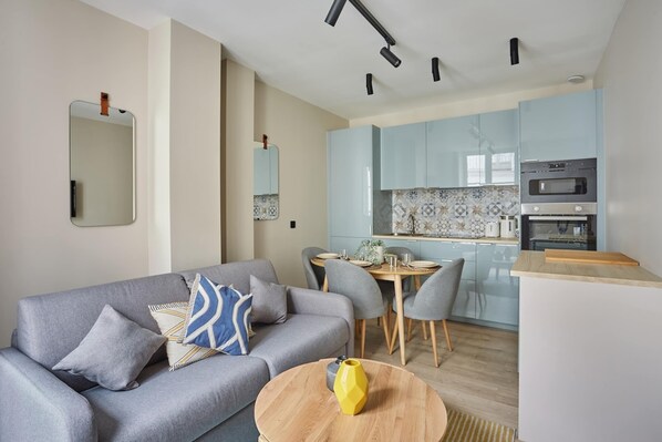 A modern living room with a grey sofa, a dining table with chairs, and a kitchen area with light blue cabinets and a built-in oven.
