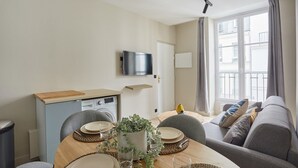 A modern living room with a dining area, a flat-screen TV mounted on the wall, a sofa, and a window with curtains. - Charming Apartment in the Center of Paris / Bourse (Paris)