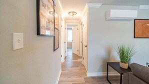 Een hal met houten vloer, witte wanden en kunstwerken in lijsten. Op een klein tafeltje staat een plant en aan de wand is een airconditioningsunit bevestigd.