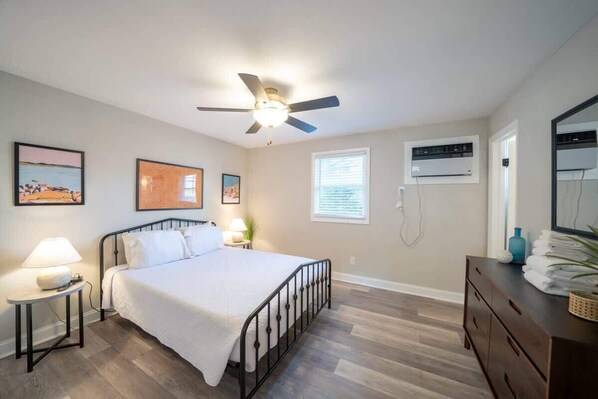 A bedroom with a bed, a ceiling fan, a bedside table with a lamp, a dresser, and a window with blinds. - OV Mosaic - Drift Suite (Norfolk)