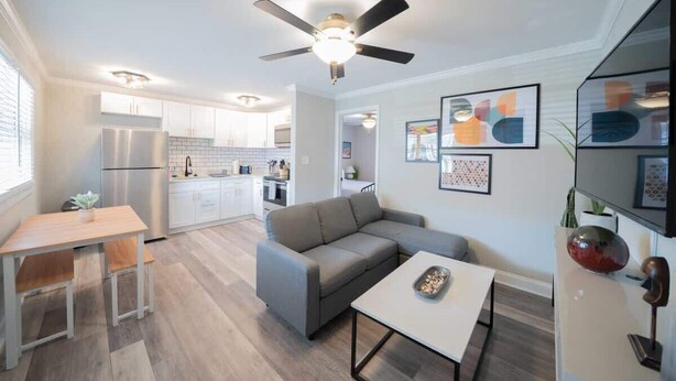A modern living room with a grey sofa, a coffee table, and a dining area with a table and chairs. There is a kitchen in the background with white cabinets and a refrigerator. - OV Mosaic - Drift Suite (Norfolk)