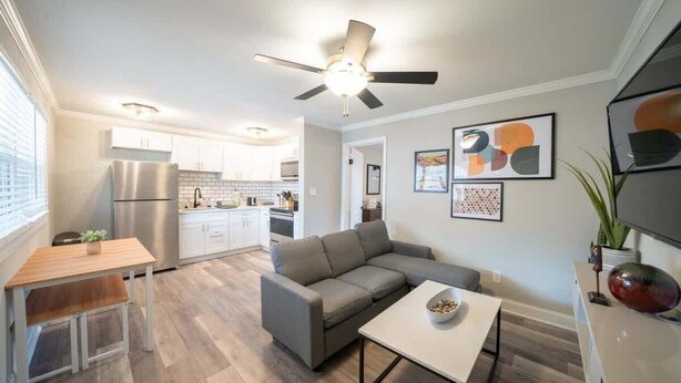 A modern kitchen with white cabinets, a stainless steel refrigerator, and a dining area with a wooden table and a gray sofa. - OV Mosaic - Summer Suite (Norfolk)
