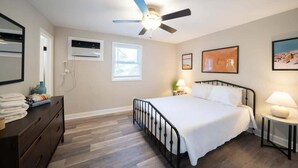 A bedroom with a bed, a ceiling fan, an air conditioning unit, a window, a bedside table with a lamp, and a dresser. - OV Mosaic - Summer Suite (Norfolk)