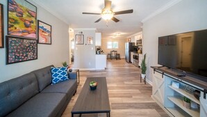 A modern living room with a sofa, coffee table, and TV. There is a kitchen in the background. - OV Mosaic - Buoy Suite (Norfolk)