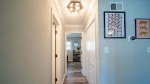 A hallway with a wooden floor, white walls, and a ceiling light fixture. There are framed pictures on the wall and a door at the end of the hallway. - OV Mosaic - Buoy Suite (Norfolk)