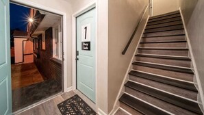A hallway with a light blue door marked "1 Suite," a wooden staircase, and a doormat. - OV Mosaic - Buoy Suite (Norfolk)