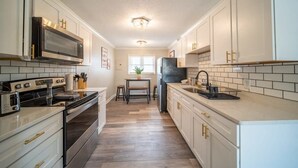 A kitchen with white cabinets, a microwave, a stove, and a refrigerator. There is a window and a door leading to another room.