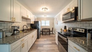 A modern kitchen with white cabinets, granite countertops, a built-in microwave, and a dining area with a table and chairs. - OV Mosaic - River Suite (Norfolk)