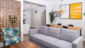 A living room with a grey sofa, a wooden partition, and a dining table with chairs. There is a ceiling fan and a clock on the wall. - Sojourn Beach Side Paradise Cove Suite (Norfolk)