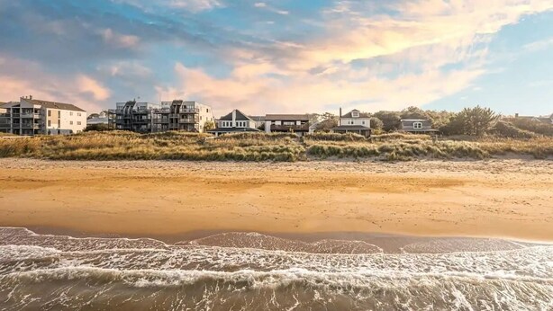 A beachfront property with multiple buildings, including a large house and several smaller structures, situated on a sandy beach with waves in the foreground. - Sojourn Beach Side Paradise Cove Suite (Norfolk)