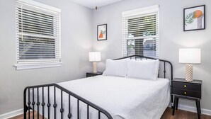 A bedroom with a bed, two bedside tables, and two windows with blinds.