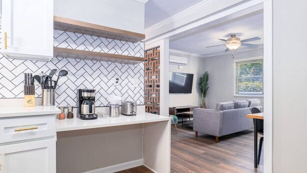 A modern kitchen with a herringbone tile backsplash, a built-in coffee maker, and a dining area with a gray sofa and a ceiling fan.