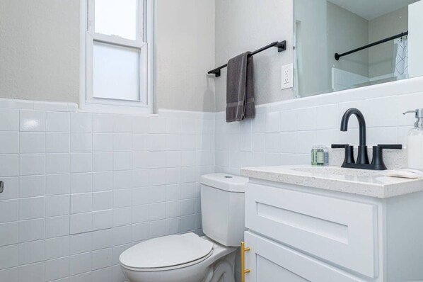Un baño moderno con azulejos blancos, inodoro blanco, grifo negro y un espejo sobre el lavamanos.