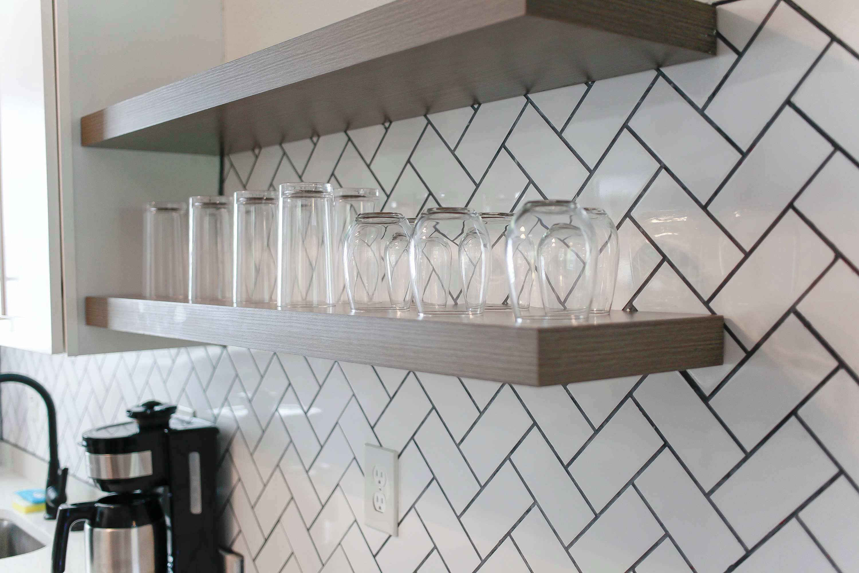 A kitchen with a herringbone patterned backsplash, a wooden shelf holding clear glasses, and a coffee maker on the counter.