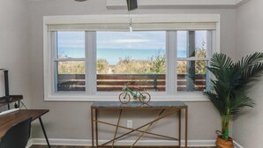 Een kamer met een groot raam met uitzicht op een strand, een plafondventilator, een tafeltje met een fiets en een bloempot.