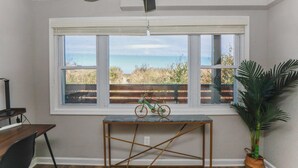 A room with a large window overlooking a beach, a ceiling fan, a small table with a bicycle, and a potted plant.