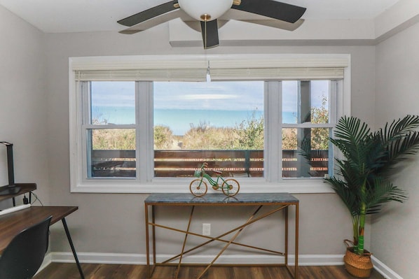A room with a large window overlooking a beach, a ceiling fan, a small table with a bicycle, and a potted plant.