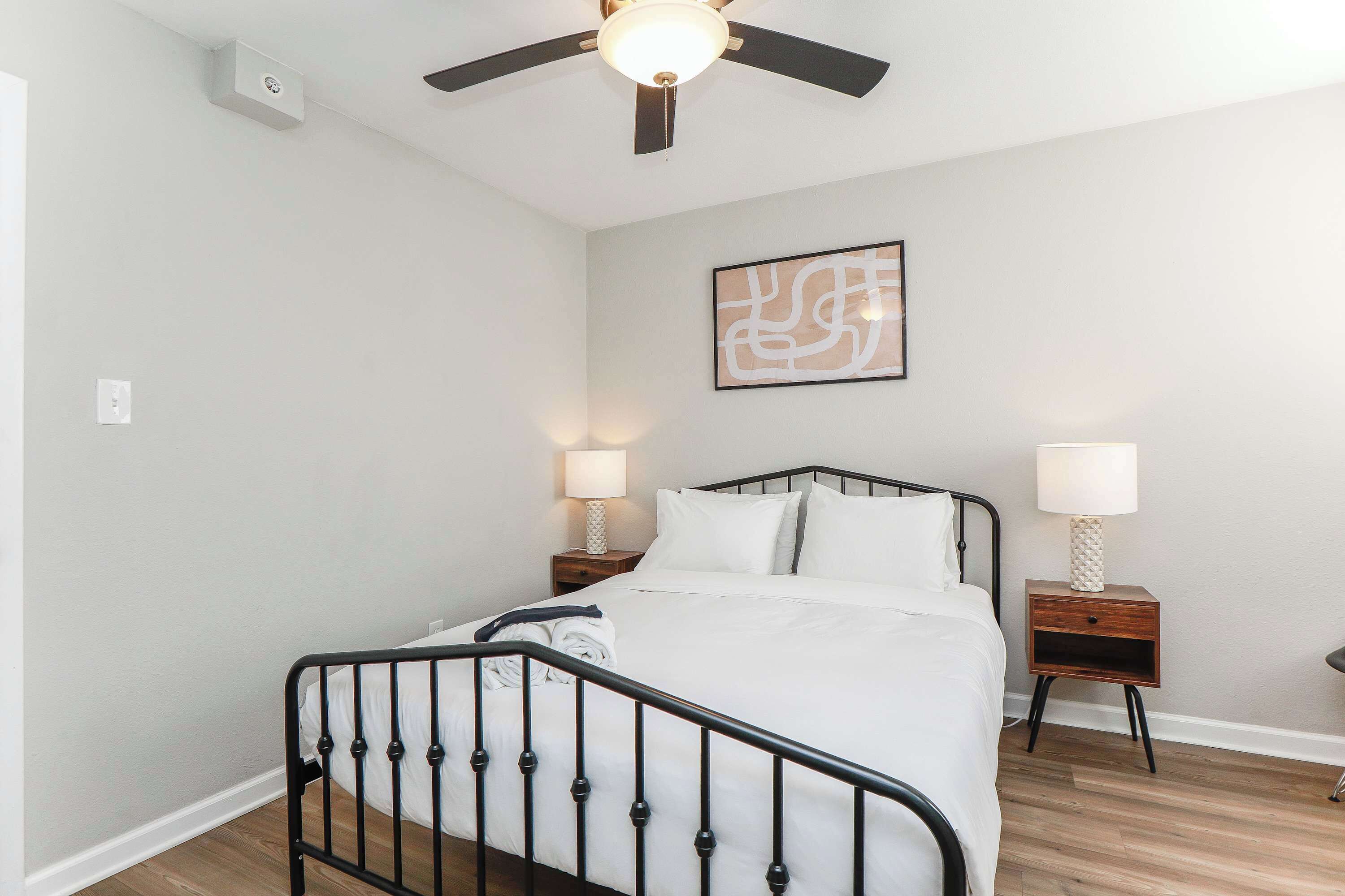 A bedroom with a bed, a ceiling fan, two bedside lamps, and a wall-mounted abstract art piece.
