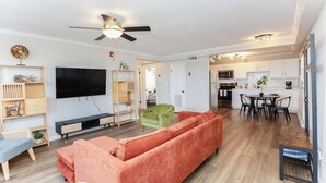 A modern living room with a large TV, a sofa, a dining table with chairs, and a kitchen in the background.