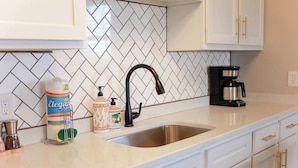 A kitchen with a stainless steel sink, white cabinets with gold handles, a herringbone-patterned backsplash, and a coffee maker on the counter.