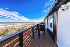 A deck with a railing overlooking a beach, a table with chairs, and a building with windows.