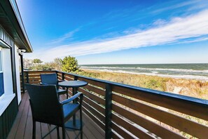 A balcony with wicker chairs and a table, overlooking a beach with dunes and the ocean.