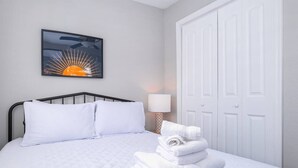 A neatly made bed with white linens, a wall-mounted artwork depicting a sun, and a stack of folded towels on the bed.