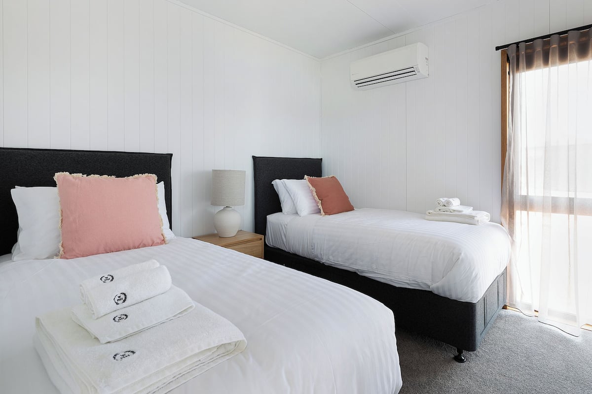 A hotel room with two beds, white bedding, a black headboard, a bedside lamp, a window with sheer curtains, and a wall-mounted air conditioner.