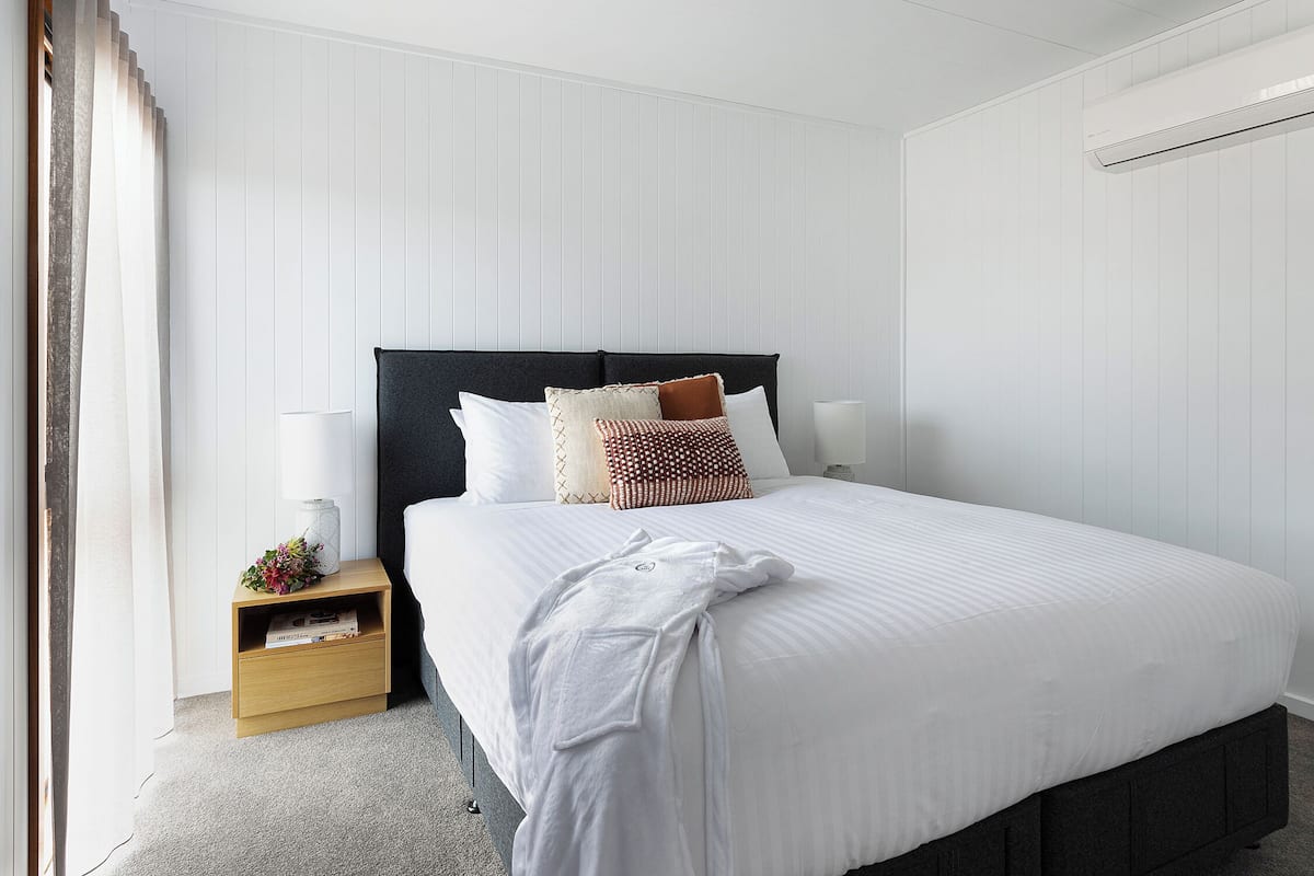 A neatly made bed with white bedding, a dark headboard, and several decorative pillows. There is a bedside table with a lamp and a small shelf with books and a vase of flowers. An air conditioning unit is mounted on the wall above the bed.