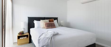Ein ordentlich bezogenes Bett mit weißer Bettwäsche, ein dunkles Kopfteil und mehrere dekorative Kissen. Ein Nachttisch mit Lampe und eine kleine Ablage mit Büchern und einer Blumenvase. Eine Klimaanlage ist an der Wand über dem Bett angebracht.