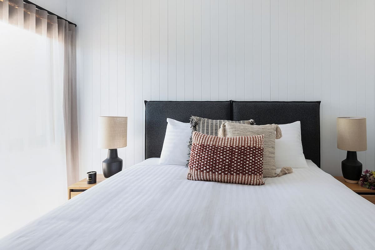 A neatly made bed with a dark headboard, two bedside lamps, and a small plant on the nightstand.