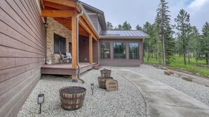 A modern house with a wooden exterior, a stone foundation, and a covered porch. There are two large barrels and a sign that reads "Hills Coop" on the gravel driveway.