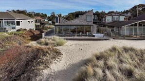 Una casa moderna frente a la playa con un porche acristalado, una playa arenosa en primer plano y otras casas al fondo.