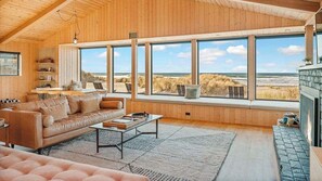 A wooden interior with a sofa, coffee table, and large windows overlooking a beach.