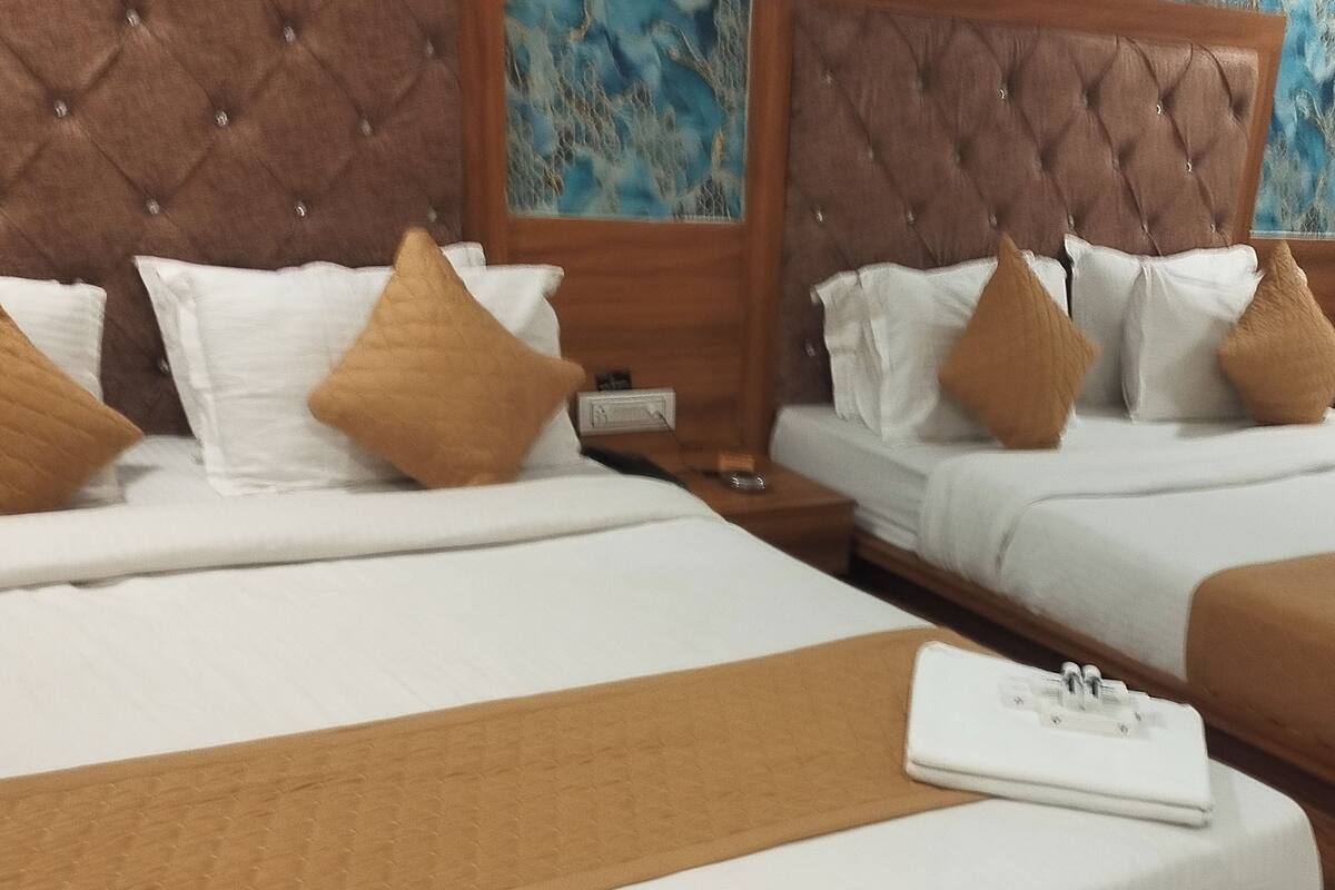 Hotellihuoneessa on kaksi sänkyä, joissa on valkoiset lakanat ja ruskeat koristetyynyt. Sängyn päädyssä on puinen koristepaneeli, jossa on kuvioitu pinta, ja sängyn vieressä on pieni yöpöytä, jossa on lamppu.