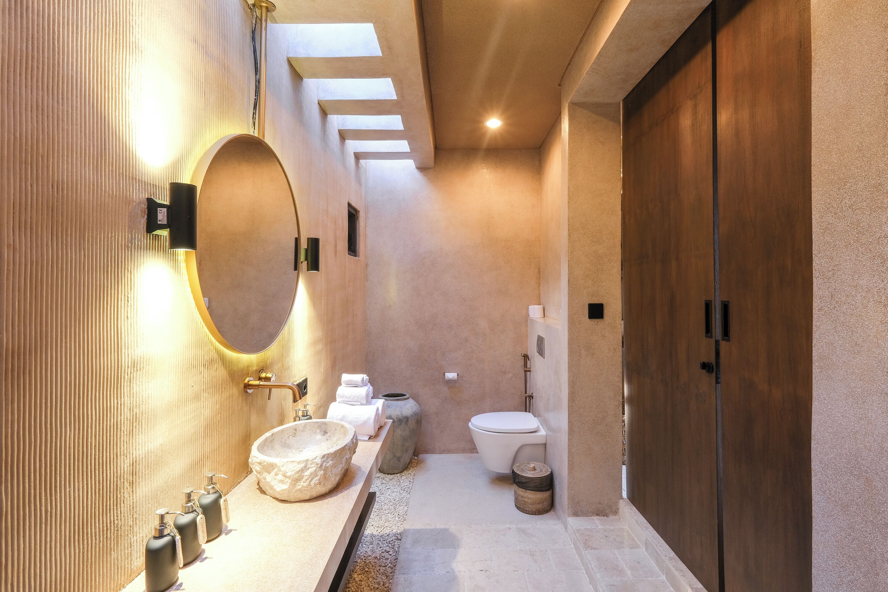 A modern bathroom with a large oval mirror, a stone sink, and a wooden door.