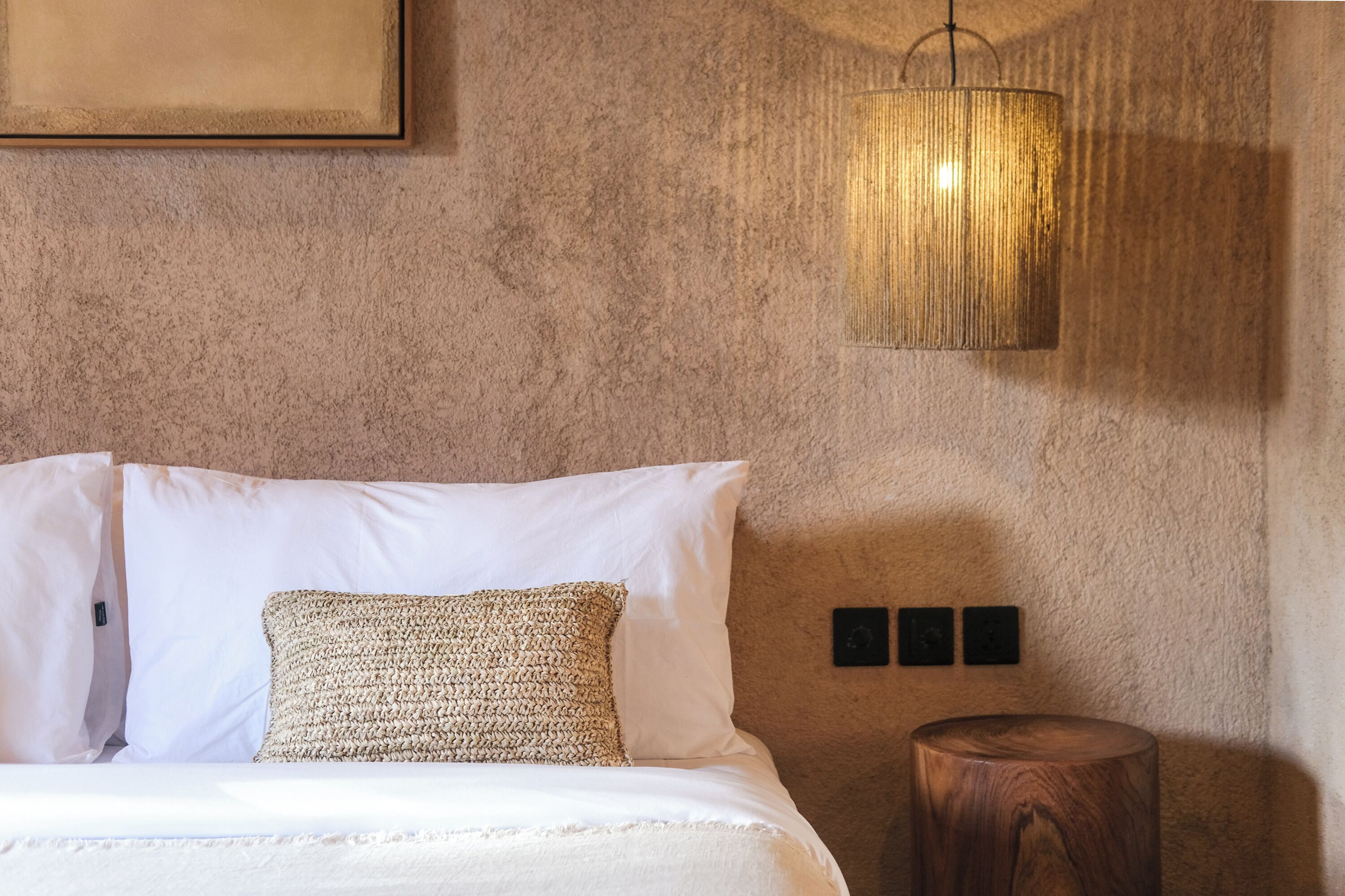 A bedroom with a bed, a textured wall, a hanging light fixture, and a wooden stool.
