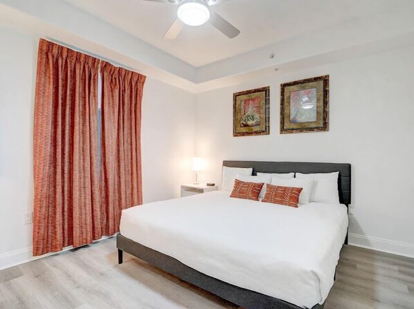 Un dormitorio con cama, cortinas, una mesita de noche y dos cuadros en la pared.