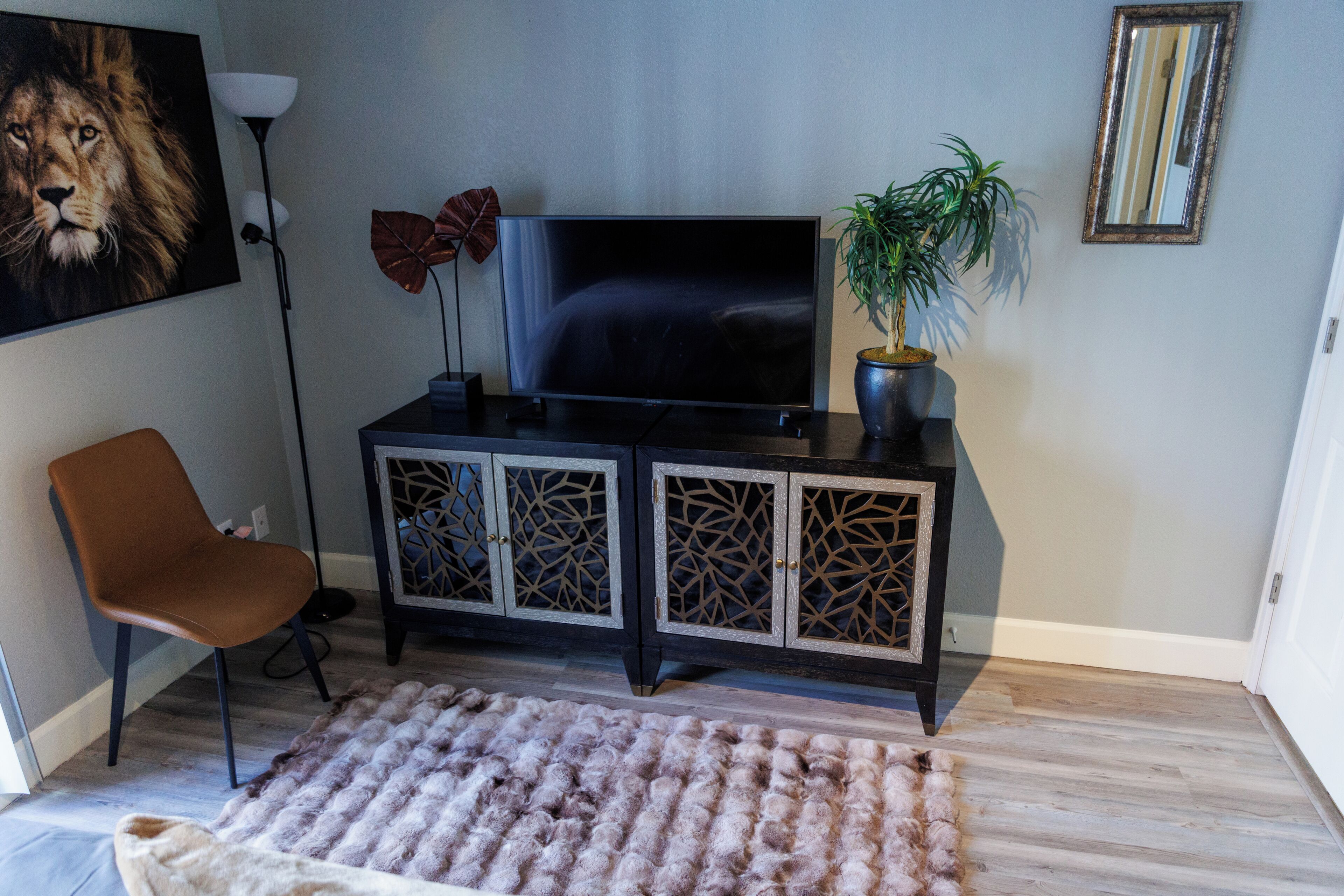Un salon avec un téléviseur posé sur un meuble noir, un fauteuil marron, un tapis à longues mèches et une plante en pot.