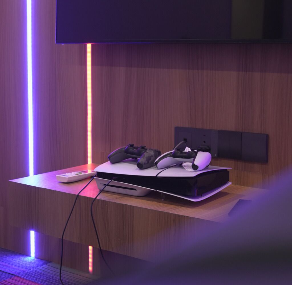 Un equipo de juego con una consola, dos mandos y un auricular sobre una mesa de madera, con un televisor montado en la pared.