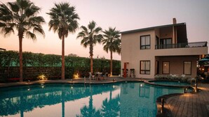 A swimming pool with a wooden deck, palm trees, and a modern building with balconies. - Villasita (Haridwar)