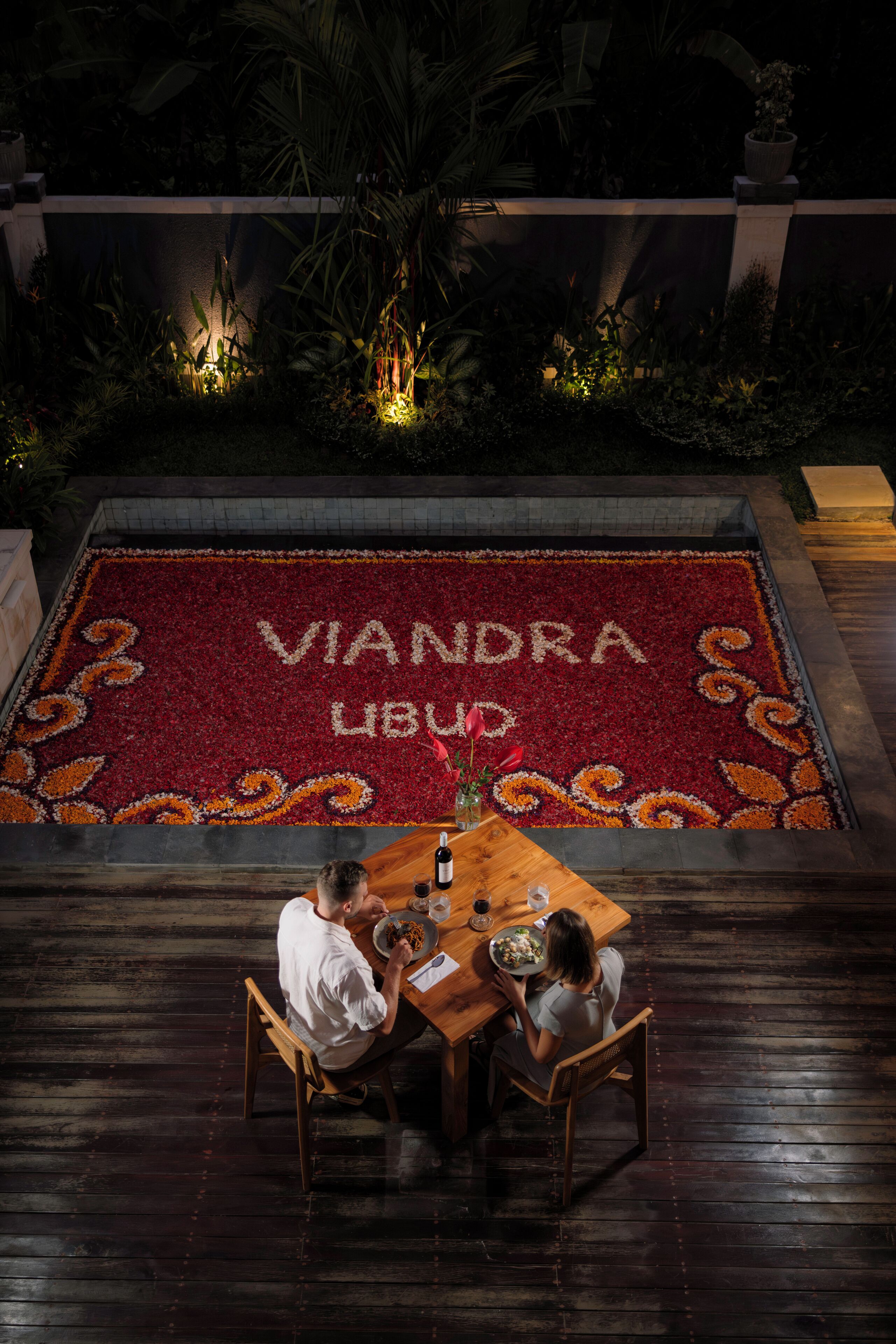 A dining area with a table and chairs, a wooden floor, and a decorative flower arrangement with the text "VIANDRA LUBU" on the ground.