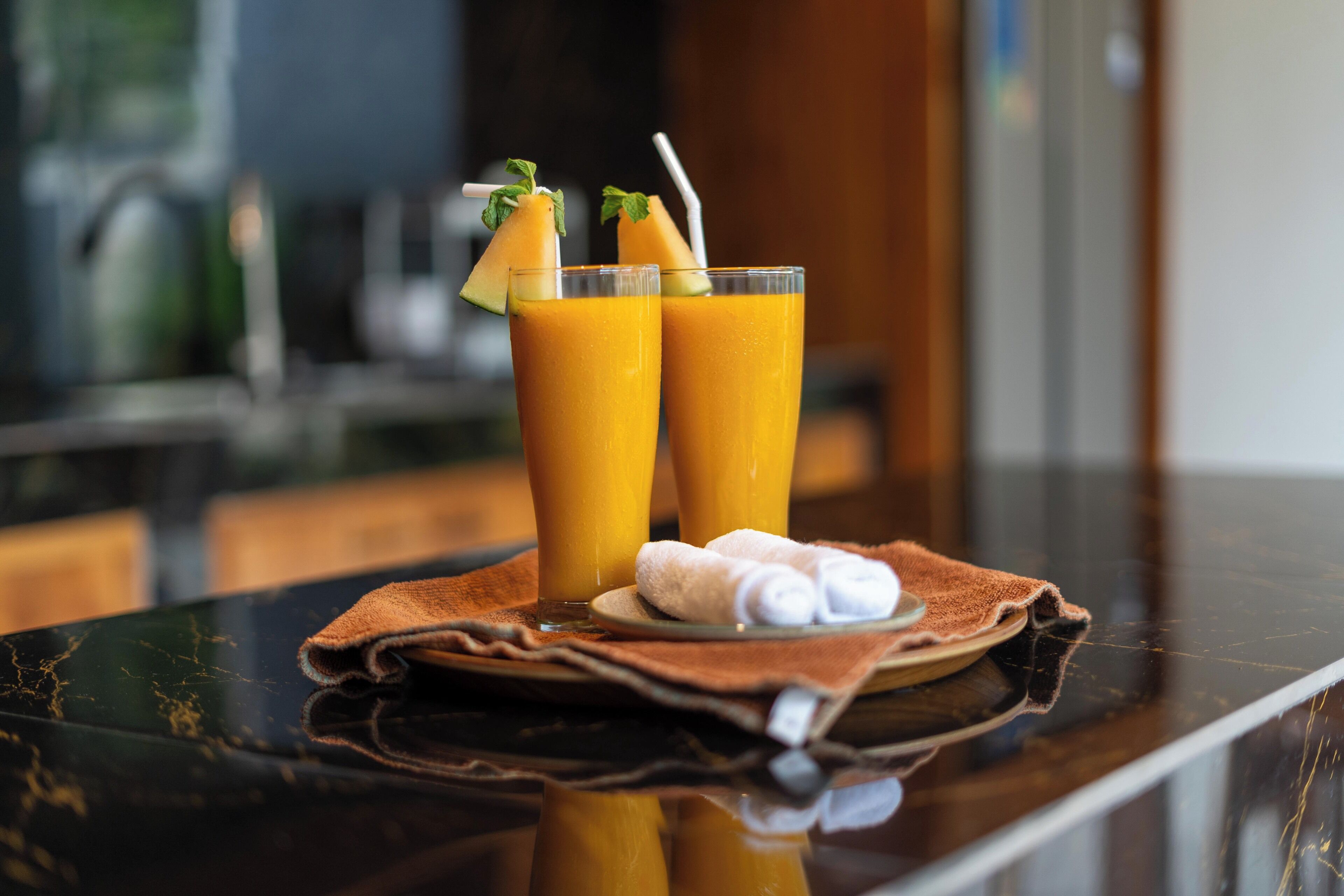 Dos vasos altos de jugo de naranja con pajitas y hojas de menta sobre una mesa negra reflectante.