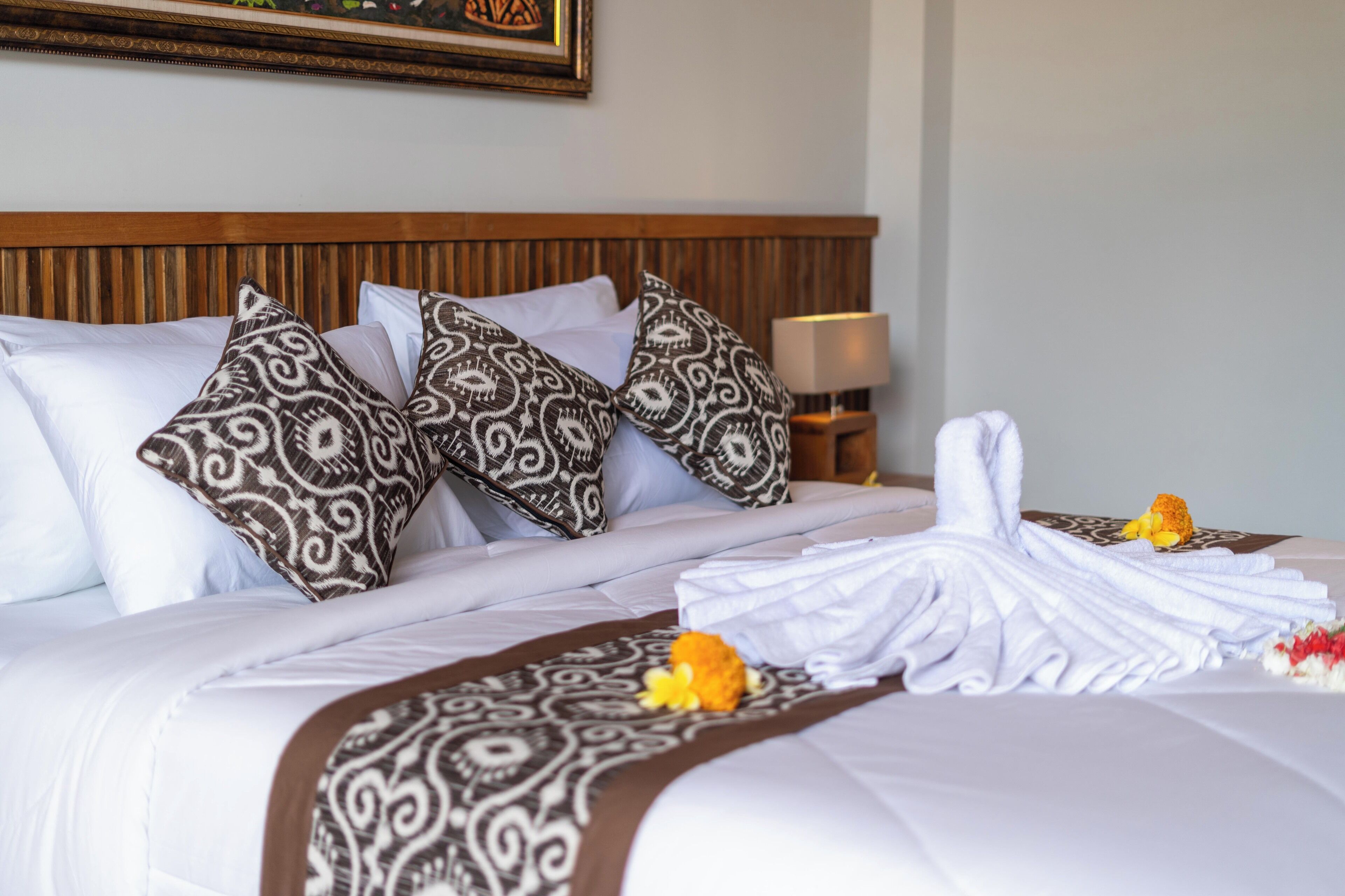 A neatly made bed with patterned pillows, a folded towel, and a flower on the bedspread.