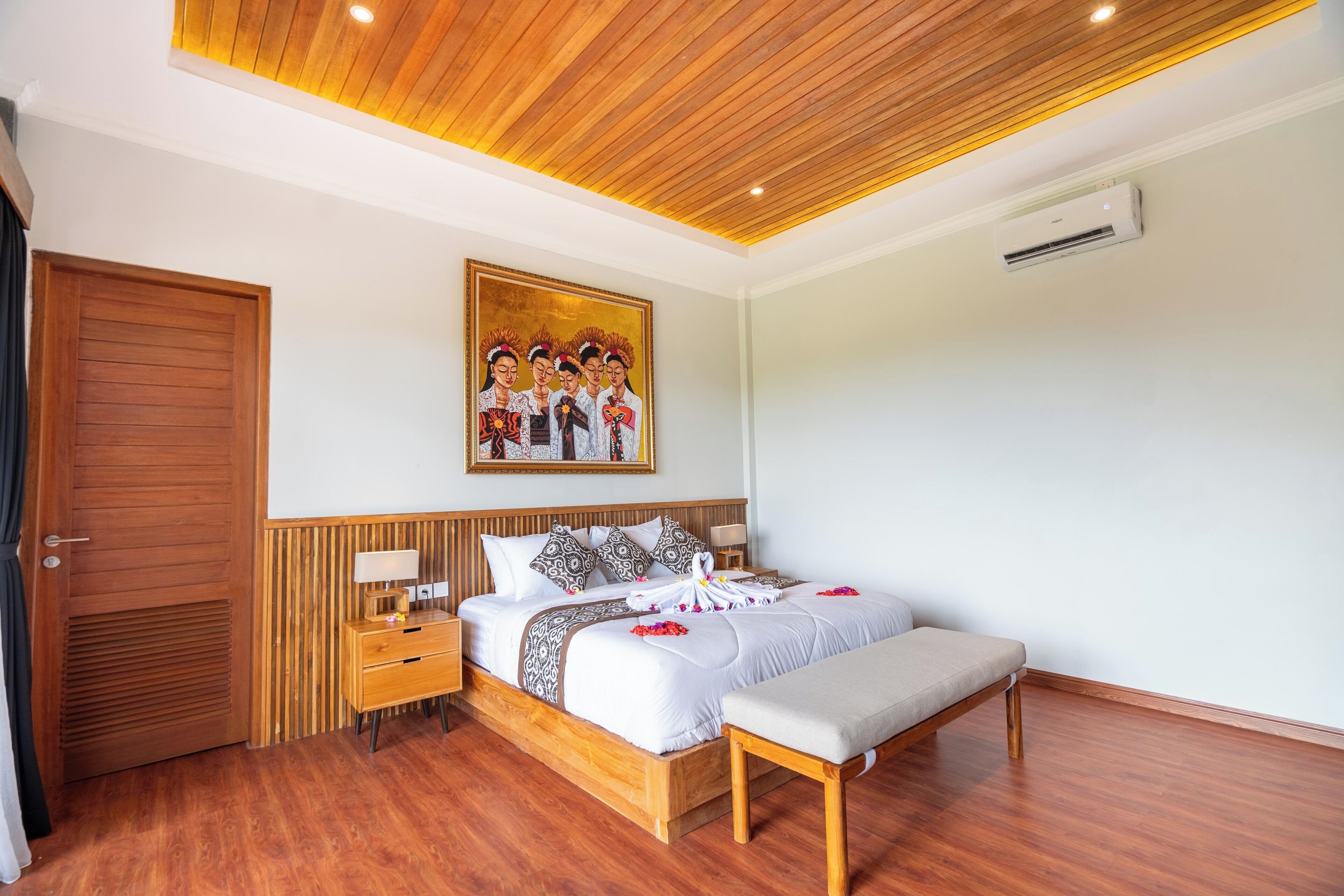 A hotel room with a wooden ceiling, a bed with white bedding, a bench, a bedside table with a lamp, and a painting on the wall.