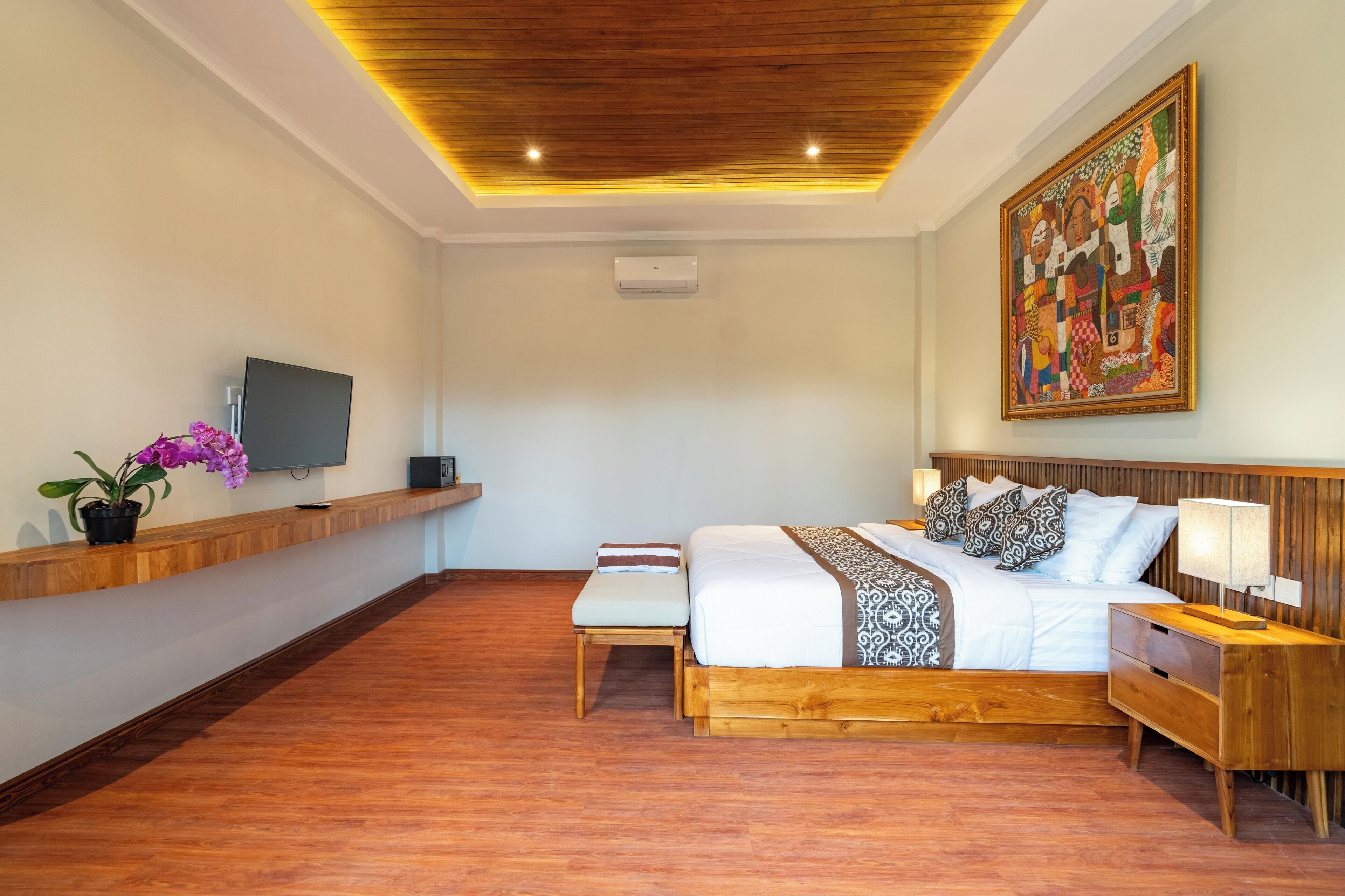 A modern hotel room with a wooden ceiling, a large bed with patterned bedding, a bedside table with a lamp, a TV on a wooden shelf, and a colorful abstract painting on the wall.